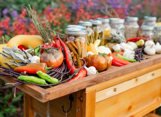 Closeup of wooden cart laden with fresh vegetables and filled mason jars, against a garden backdrop of red and purple blossoms.