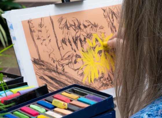 A close-up view over the shoulder of an individual using a bright yellow pastel stick to draw flowers on orange paper, with a box of assorted colored pastels in the foreground.