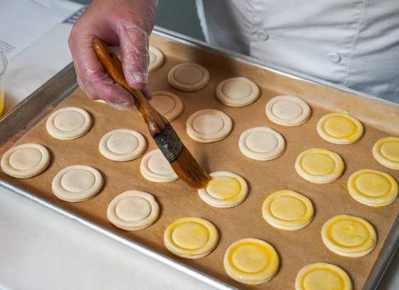 A baker wearing plastic gloves uses a pastry brush to apply a golden egg wash onto rows of small, circular, unbaked pastries arranged on a parchment-lined baking sheet.