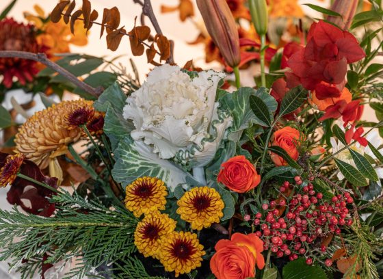 A close-up of a decorative floral arrangement featuring a central white ornamental cabbage, surrounded by warm-toned orange and yellow chrysanthemums, red berries, and autumn-colored leaves.