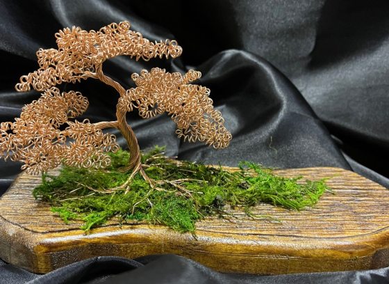 A wire bonsai sculpture on a wooden board sitting against a black fabric backdrop.