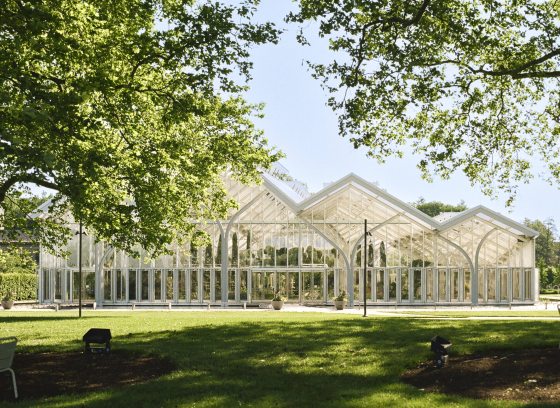 Large trees shade a green lawn in front of a large glass house with peaked arches.