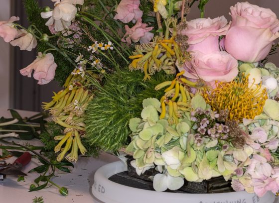 A floral design in process, featuring pink roses, goldenrod accents, and other floral material, in a shallow white vase.