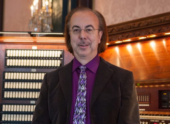 a person dressed in a dark suit, purple shirt, and tie, faces the camera  with his back to a large organ console