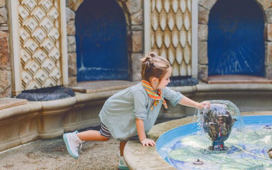 A small child stands on one foot as they reach their hand on to a stone water fountain