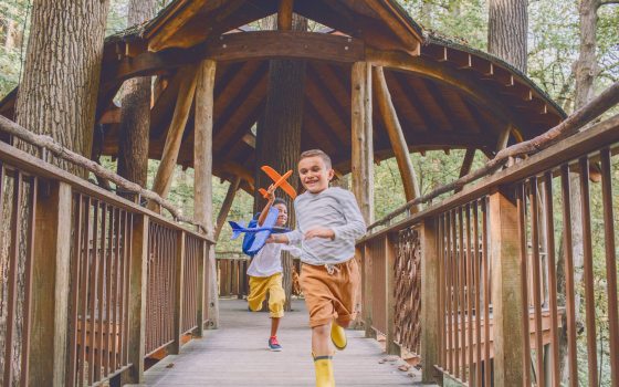 Two children holding toy airplanes run along a wooden walkway out of a wooden treehouse
