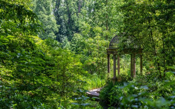A stone gazebo sits beside a small lake tucked into a forest of bright green trees