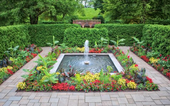 a square fountain surrounded by flower beds in bloom and enclosed by a green hedge