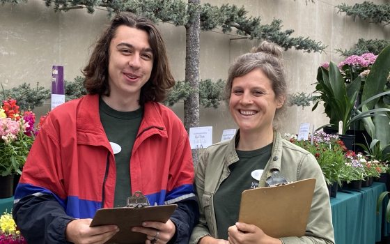 Two students with clipboards smile at camera during a plant sale