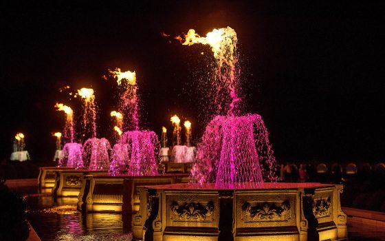 A row of octagonal stone basins at night, with fountains in basketweave formation, lit up in fuchsia and topped by fire.
