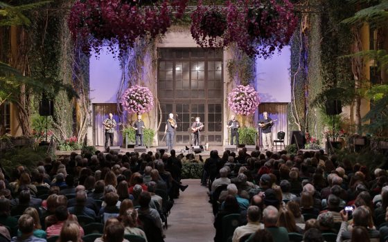 performers on a stage in an indoor garden setting, with audience in foreground