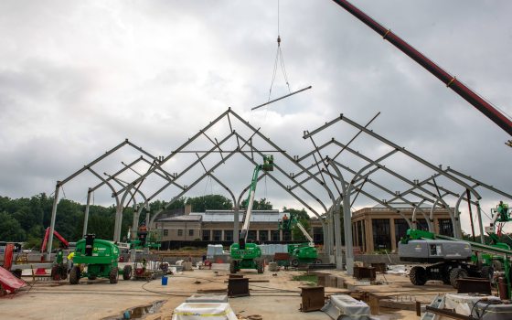 Three rows of structural beams, pyramidal in shape, being assembled by a large crane and four workers in green JLG brand lifts
