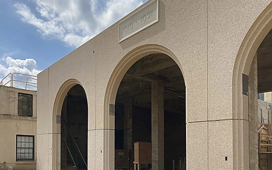 angled shot of facade with stone archways, under construction, with datestone above one arch