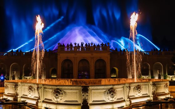 backlit images of people standing on a stone balcony watching and photographing blue fountains against a night sky, flanked by 2 foreground fountain jets topped by flames