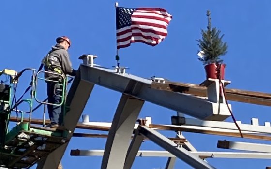 a construction worker stands in a bucket lift at the top of arched steel beams, decorated by an American flag and a Christmas tree