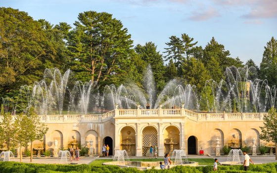 Fountains shoot up in arched forms over a white stone facade in a garden during the day