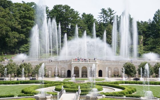 Fountains shoot water high into the air during the day over carved white stone structure and green boxwoods