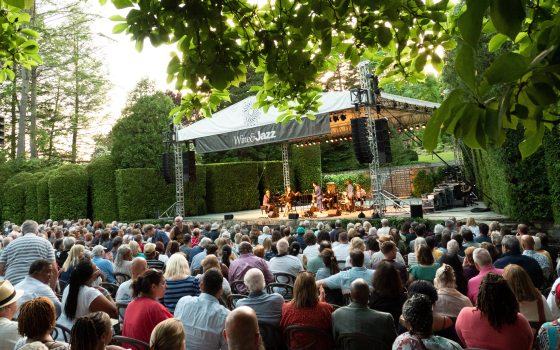 an audience watches musicians playing on an outdoor stage amid green trees