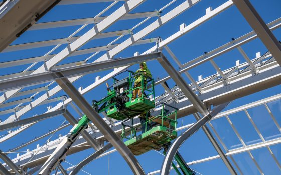 a construction worker atop a large crane is framed by steel arches against a backdrop of blue sky