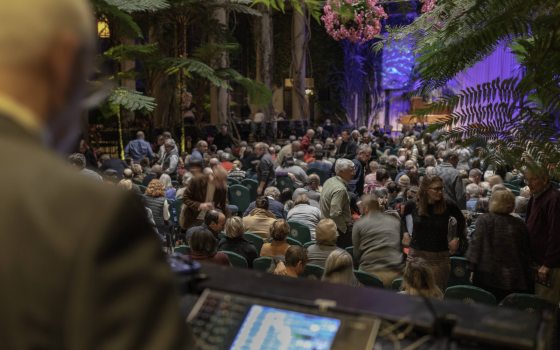Someone looks at a computer control panel in the foreground of an indoor garden transformed into a performance hall, where an audience awaits.