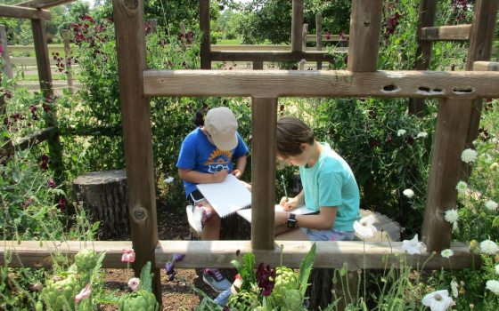 Two young students sitting inside a wooden structure drawing on notepads.