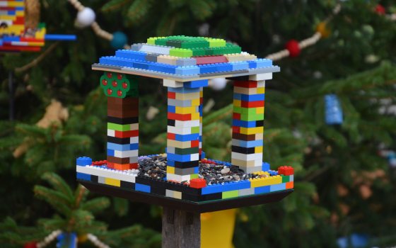A birdhouse made from lego blocks hanging on an outdoor evergreen tree.