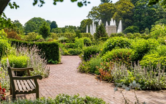 A brick walkway with a wooden bench winds through green garden beds with fountains in the distance.