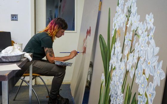 A person in an art studio hunched over painting flowers on a large-scale white board.