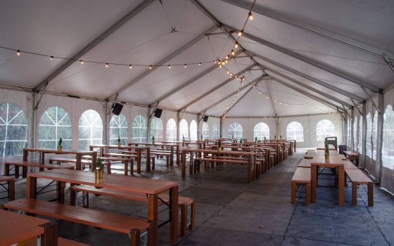 Inside shot of white outdoor dining tent with arched plastic windows, with wood benches and chairs set up on outdoor flooring, and small white light bulbs strung overhead.