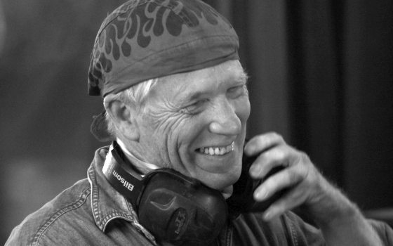 Black & white portrait of person smiling broadly, wearing a headscarf and denim shirt, with headphones around their neck.