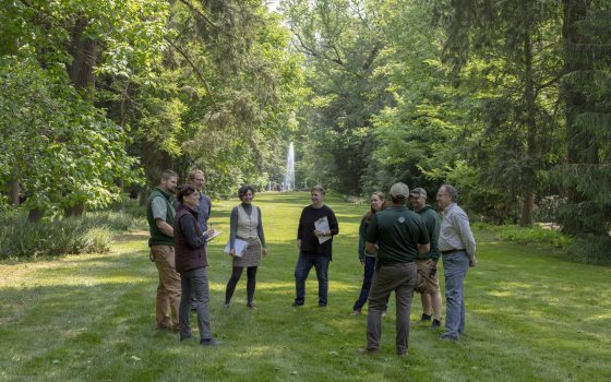 Nine members of Longwood's science team, two carrying clipboards, and one carrying a journal and pen, seem to enjoy a lighthearted moment on a grassy lawn amid tall trees, with a fountain and guests in the distant background.