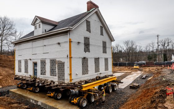An old white house being moved on a large trailer with wheels.
