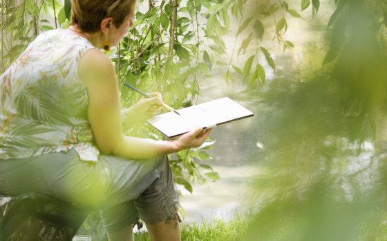 A person sits with a handheld easel and paintbrush amid the greenery of a weeping tree in a garden.