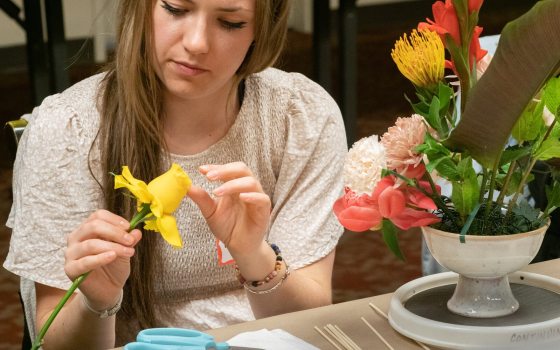 A seated person creating a floral arrangemetn, holding a yellow rose.