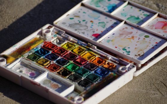 A used box of watercolor paints, opened and laying on a tan surface.