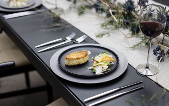 Closeup of a long narrow black table positioned diagonally acroos the frame, with a black plate atop a silver server at bottom right, set with flatware, a glass of red wine, and displaying a souffle-like item and salad atop the plate.