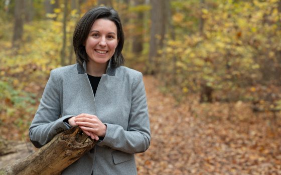 A woman with black hair in a grey jacket stands in a forest and smiles for a photo