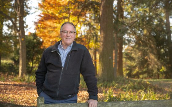 A man with glasses in a zip up sweater poses amongst bright fall foliage 