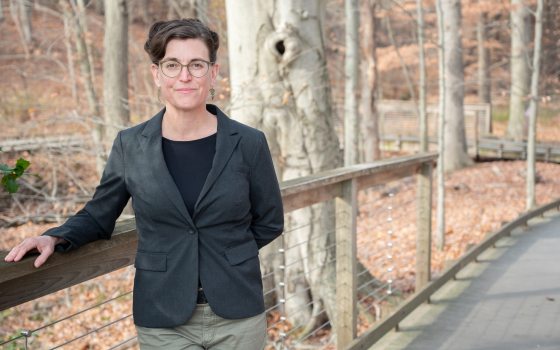 A woman with black hair and glasses with a black blazer rests her arm on a railing on a boardwalk in a forest. 