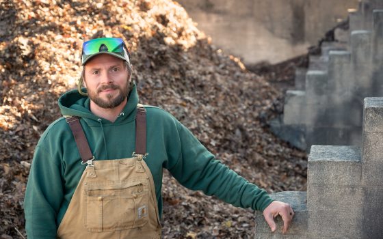 A man in a green sweatshirt and overalls stands near a compost pile.