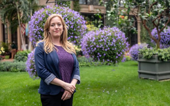 A woman with blonde hair poses for a portrait amid hanging baskets of purple plants.