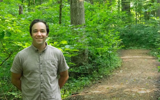 A tan man with thick black hair and a gray button down shirt smiles for a photo in a woodland setting.