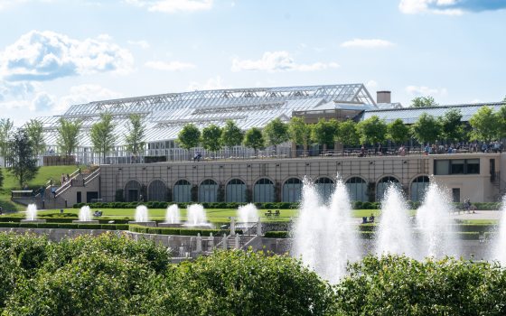 Fountains can be seen in the forefront of the photo. In the background, a new glass conservatory can be seen on top of an arched lower façade and next to another building.  