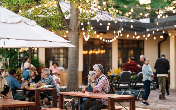 People eat and drink at umbrella-covered tables under strings of lights in an outdoor setting.