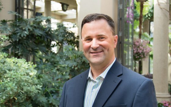 Man with short dark hair wearing blue suit jacket over a light blue button up shirt smiling at the camera