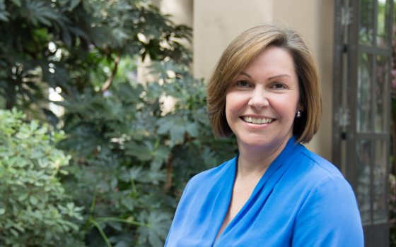A person in a blue shirt, with shoulder length brown hair, smiling at the camera.