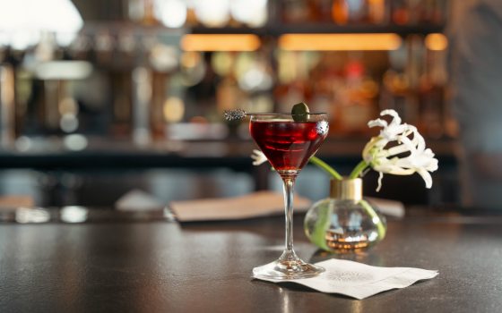 Closeup of a martini glass filled with a red drink, topped by a skewered olive, resting atop a white napkin bearing the Longwood logo, backed by a white flower in a small glass vase.