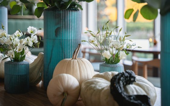 Closeup of seasonal restaurant decor, including white pumpkins, a green gourd, and cylindrical turquoise vases filled with white flowers and green foliage.