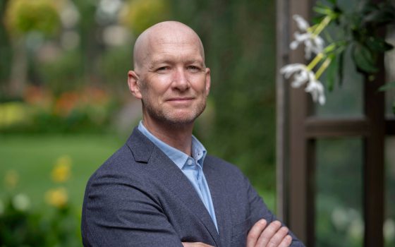 A headshot of a white man in a suit jacket with blue dress shirt with greenery in the background 