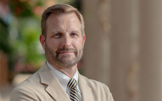 Man with facial hair in light suit with dark striped tie looking at camera